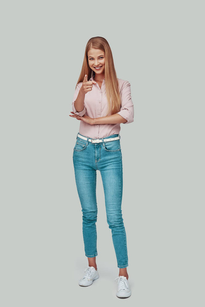 Piena lunghezza di attraente giovane donna guardando la fotocamera e indicando mentre in piedi contro sfondo grigio
 - Foto, immagini