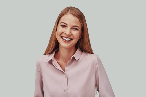 Attractive young woman looking at camera and smiling while standing against grey background - Zdjęcie, obraz