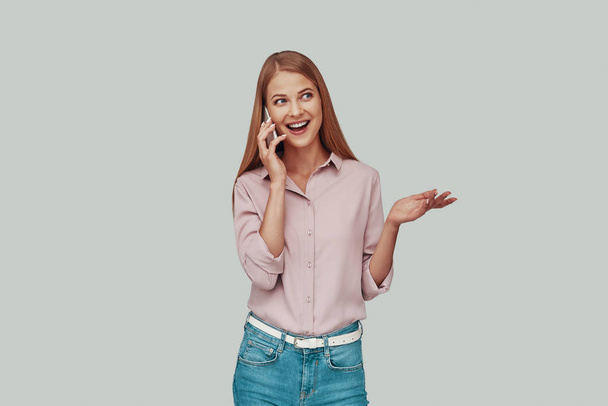 Vonzó fiatal nő beszél az okos telefont, és mosolyogva állva ellen szürke háttér - Fotó, kép