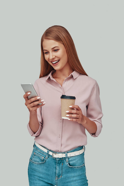 Attractive young woman using smart phone and smiling while standing against grey background - Foto, Imagem