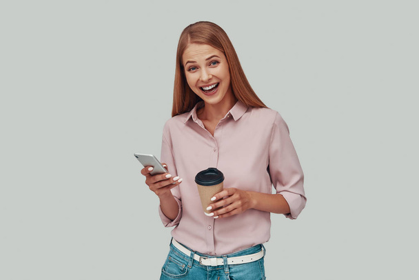 Attraente giovane donna utilizzando smartphone e sorridente mentre in piedi su sfondo grigio - Foto, immagini
