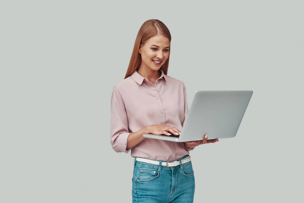 Mujer joven atractiva usando el ordenador portátil y sonriendo mientras está de pie sobre fondo gris
 - Foto, Imagen