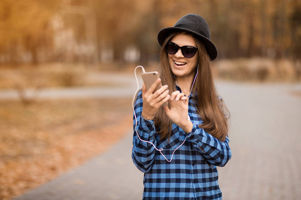 Jovem mulher de beleza em camisa azul chapéu preto e óculos escuros escrevendo mensagem no celular enquanto caminhava pela rua de outono
 - Foto, Imagem