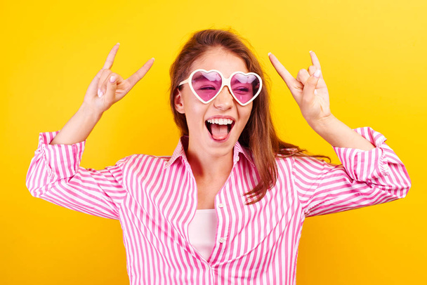 Yeah, feeling cool and awesome. Excited happy funny young  woman lifts mood from loud music, shows rock n roll gesture, in glasses in the shape of a heart on a yellow bright background - Foto, immagini