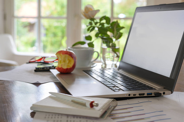 Thuiskantoor werkplek voor een klein bedrijf, op het Bureau een laptop met leeg scherm, papieren, koffiekop, glazen en een gebeten appel - Foto, afbeelding