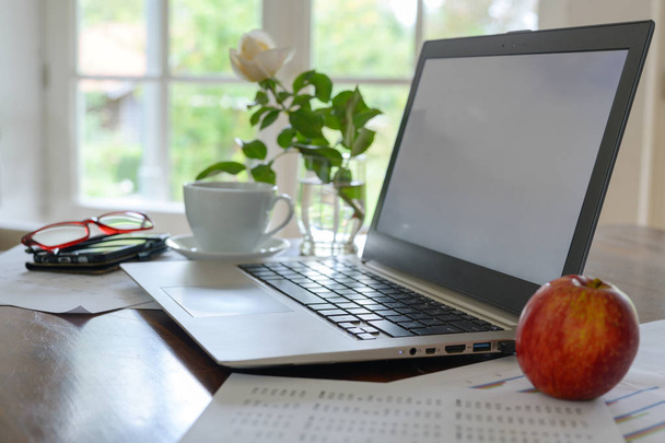 Laptop mit leerem Bildschirm, Papieren, Kaffeetasse, Gläsern und einem frischen Apfel auf einem Arbeitsplatz im Home Office am Fenster - Foto, Bild