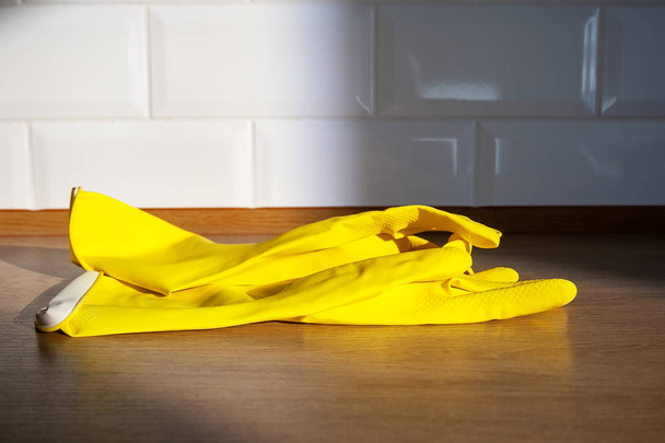 Gele rubberen handschoenen voor huishoudelijk werk liggen op een houten aanblad tegen een vervaagde muur van witte tegels op een zonnige dag - Foto, afbeelding