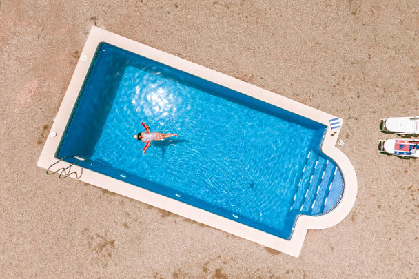 Zenith luchtfoto van een zwembad in de zomer. Jong meisje in een badpak en hoed drijvend met zwembad noedels. - Foto, afbeelding