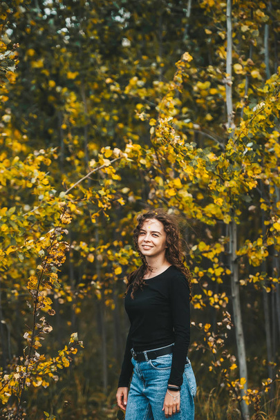 Красива стильна дівчина в чорній сорочці і в блакитних джинсах стоїть і позує в осінньому парку
. - Фото, зображення