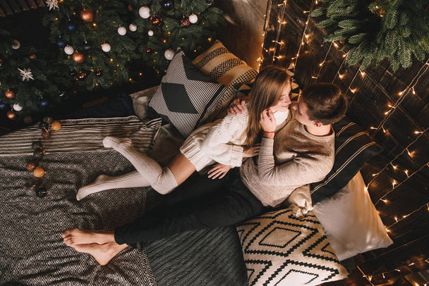 Couple on the bed in the bedroom. Dark interior. New Year's and Christmas. Hugs and kisses. Love. White sweater and high socks. Romantic meeting. A date lovers. - Фото, изображение