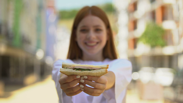 Joven hembra alegre mostrando sándwich fresco en la cámara y merienda sonriente y saludable
 - Metraje, vídeo
