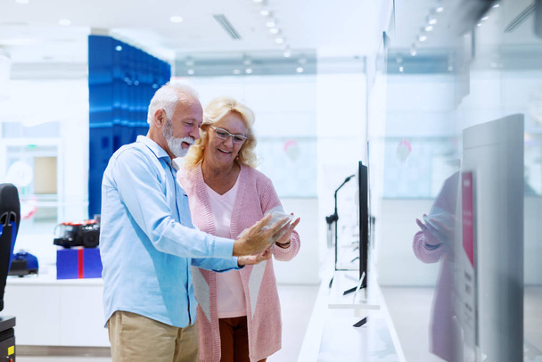 Cute uśmiechnięty kaukaski starszy para stojący w tech Store i patrząc na ceny dla TV chcą kupić. - Zdjęcie, obraz