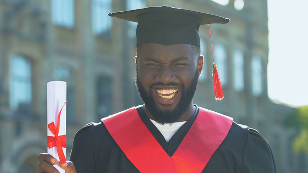 Cheerful black male student in gown holding diploma and making yes gesture - Footage, Video