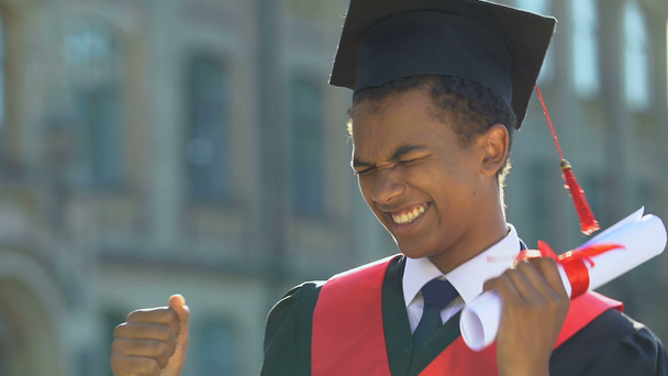 Funny afro-american boy in gown holding diploma and making dancing movements - Video