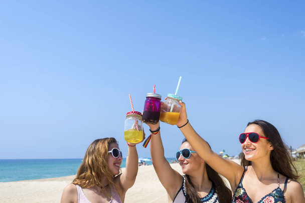 Gruppe fröhlicher junger Frauen oder Freundinnen, die am Sommerstrand auf alkoholfreie Getränke anstoßen - Foto, Bild