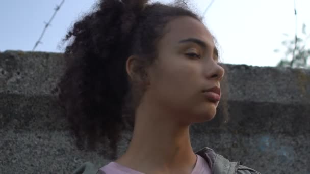 Upset African-American girl with bag standing behind wall with barber wire - Filmagem, Vídeo