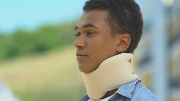 Afro-american boy in foam cervical collar looking upset outdoors, neck injuries - Séquence, vidéo