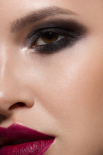 Close-up of the beauty of a woman's face with evening make-up emphasizing her expressive facial features. Smoky eyes, marsala-colored lips and clear skin. On an isolated black background - Фото, изображение