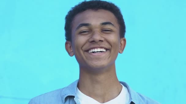 Positive African-American teen boy laughing at camera against blue background - Кадри, відео