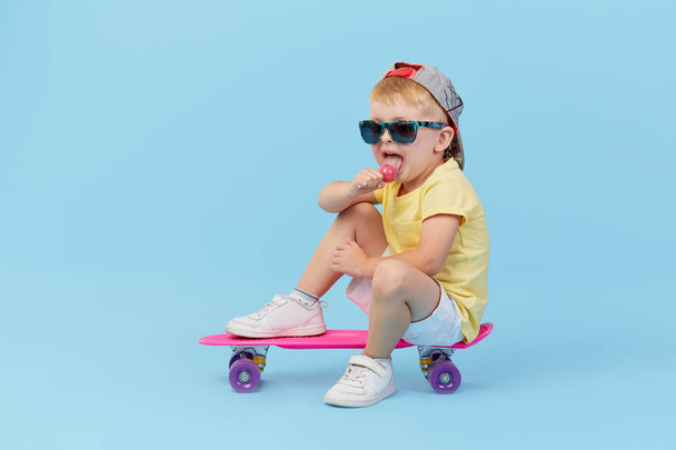 Stylish little toddler child boy in glasses with skateboard having fun over blue background - Photo, Image