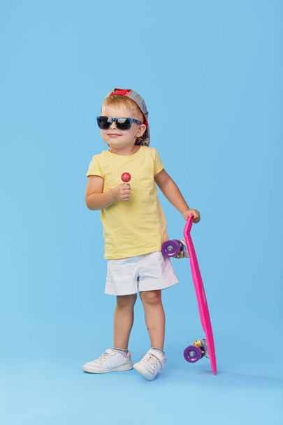 Stylish little toddler child boy in glasses with skateboard having fun over blue background - Foto, imagen