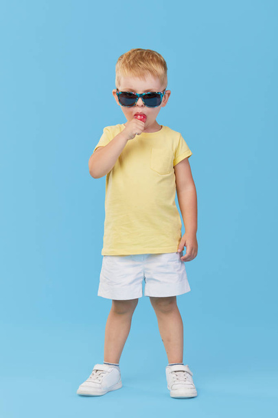 Little serious fashionable toddler boy in summer clothes and trendy sunglasses. Eats sweet candy on stick Isolated on blue background - Fotografie, Obrázek
