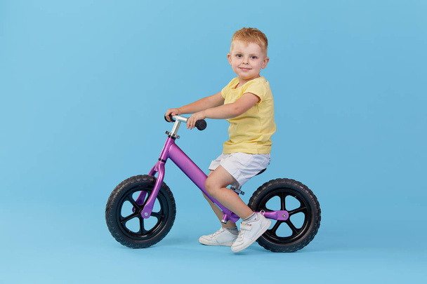 Little child boy learns to keep balance while riding a bicycle. Stylish child in yellow T-shirt on blue background - Фото, зображення
