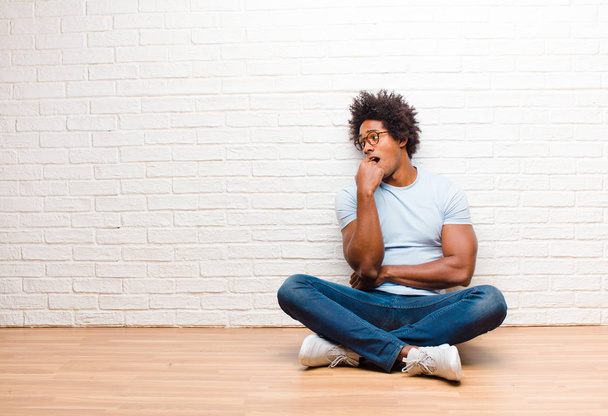 young black man with surprised, nervous, worried or frightened look, looking to the side towards copy space sitting on the floor at home - 写真・画像