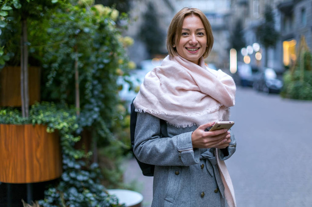 Travel blogger. Beautiful woman in a light coat and a light pink scarf with a mobile phone while walking along a city street. - 写真・画像