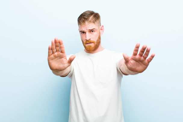 young red head man looking serious, unhappy, angry and displeased forbidding entry or saying stop with both open palms against soft blue background - Φωτογραφία, εικόνα