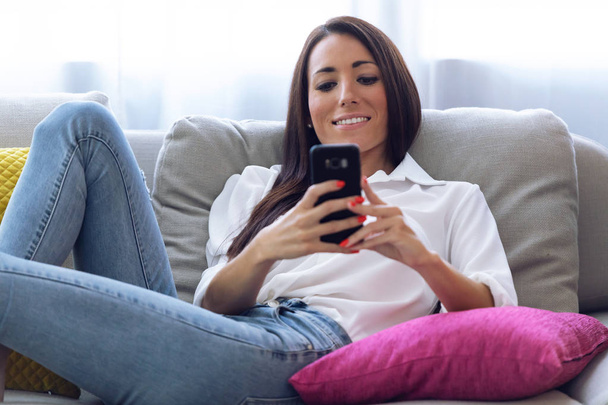 Pretty young woman using her mobile phone while sitting on sofa at home. - Фото, зображення