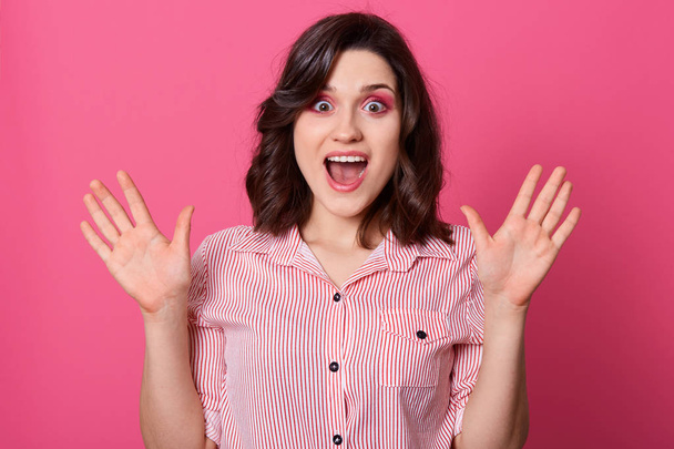 Horizontal shot of beautiful caucasian woman with shocked facial expression, keeps hands up, having widely opened eyes and mouth, has dark hair, wearing casual shirt, posing against pink wall. - Photo, Image