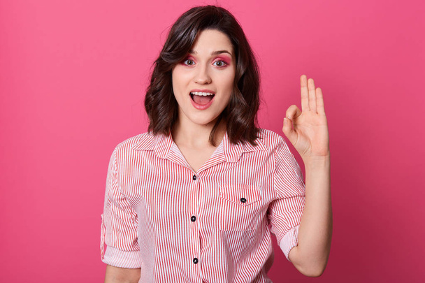 Close up portrait of beautiful young dark haired woman wearing striped shirt showing ok hand sign, keeps mouth opened and looking directly at camera, looks happy, model posing over pink background. - Foto, immagini