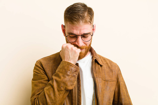young red head man feeling serious, thoughtful and concerned, staring sideways with hand pressed against chin against beige background - 写真・画像