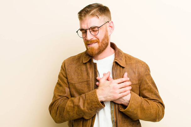 young red head man looking sad, hurt and heartbroken, holding both hands close to heart, crying and feeling depressed against beige background - Foto, imagen