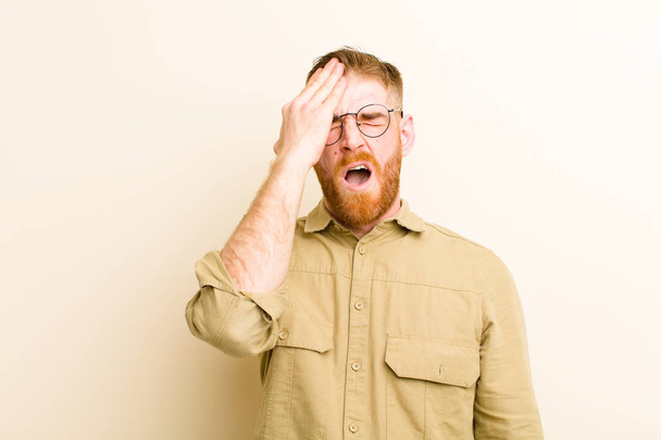 young red head man looking sleepy, bored and yawning, with a headache and one hand covering half the face against beige background - Foto, afbeelding