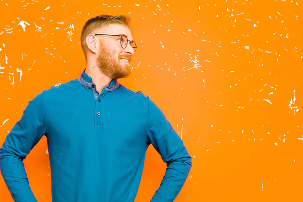 young red head man looking happy, cheerful and confident, smiling proudly and looking to side with both hands on hips against grunge orange wall - Фото, изображение