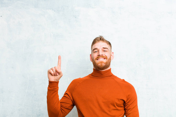 young red head man wearing turtle neck smiling cheerfully and happily, pointing upwards with one hand to copy space against concrete wall - Foto, Bild