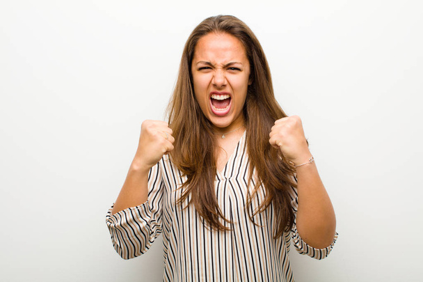 young woman shouting aggressively with annoyed, frustrated, angry look and tight fists, feeling furious against white background - Photo, image