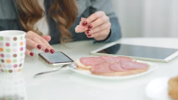 Closeup female hands scrolling mobile phone while eating sandwich - Materiał filmowy, wideo