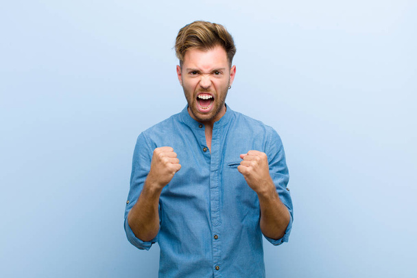 young businessman shouting aggressively with annoyed, frustrated, angry look and tight fists, feeling furious against blue background - Foto, Imagen