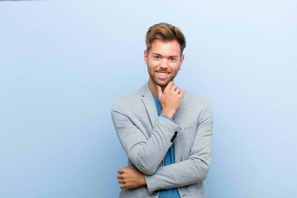 young businessman smiling, enjoying life, feeling happy, friendly, satisfied and carefree with hand on chin against blue background - Valokuva, kuva