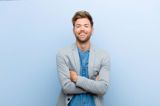 jonge zakenman op zoek als een gelukkige, trotse en tevreden strever glimlachend met armen gekruist tegen een blauwe achtergrond - Foto, afbeelding