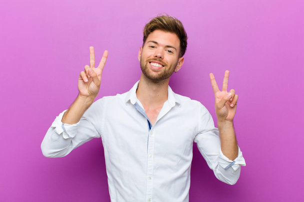 young man smiling and looking happy, friendly and satisfied, gesturing victory or peace with both hands against purple background - Photo, image