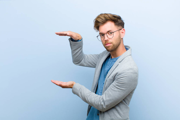 young businessman holding an object with both hands on side copy space, showing, offering or advertising an object against blue background - Фото, зображення