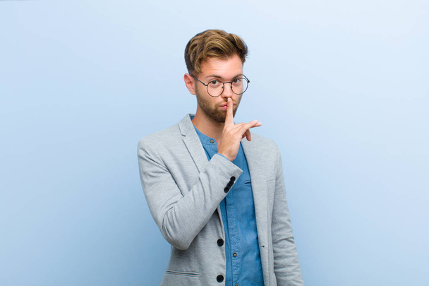 young businessman asking for silence and quiet, gesturing with finger in front of mouth, saying shh or keeping a secret against blue background - Foto, afbeelding