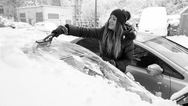 Imagem em preto e branco da senhora limpar o carro coberto de neve por escova após nevasca. Raspando o capô e janelas laterais e espelhos de neve usando o raspador e escova no início da manhã de inverno nevado
 - Foto, Imagem