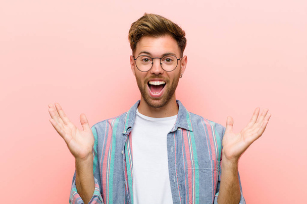 young man looking happy and excited, shocked with an unexpected surprise with both hands open next to face against pink background - Fotó, kép