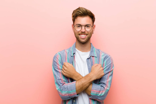 young man smiling cheerfully and celebrating, with fists clenched and arms crossed, feeling happy and positive against pink background - Photo, image