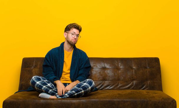 young man wearing pajamas with a goofy, crazy, surprised expression, puffing cheeks, feeling stuffed, fat and full of food . sitting on a sofa - Fotoğraf, Görsel
